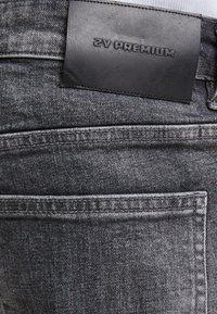 Jeans de mezclilla gris con un tejido texturizado, que presentan una etiqueta de cuero negro estampada con "ZY PREMIUM" y detalles de costura visibles.