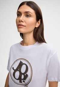 Witte katoen t-shirt met een bold bruin graphic met uitgebreide details. Ronde halslijn en korte mouwen. Eenvoudig en strak ontwerp.