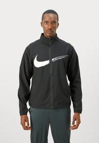 Sort Nike-jakke med fuld lynlås foran, høj krave og et fremtrædende hvidt swoosh-logo på brystet. Lavet af letvægtsmateriale.