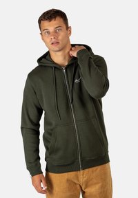 Sudadera con cremallera de color verde oscuro, con capucha ajustable, bolsillos frontales, puños de canalé y un sutil logo en el pecho, combinada con pantalones de pana beige.