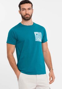 Teal cotton t-shirt with a crew neck, short sleeves, and a light blue camo-patterned patch pocket on the left side.