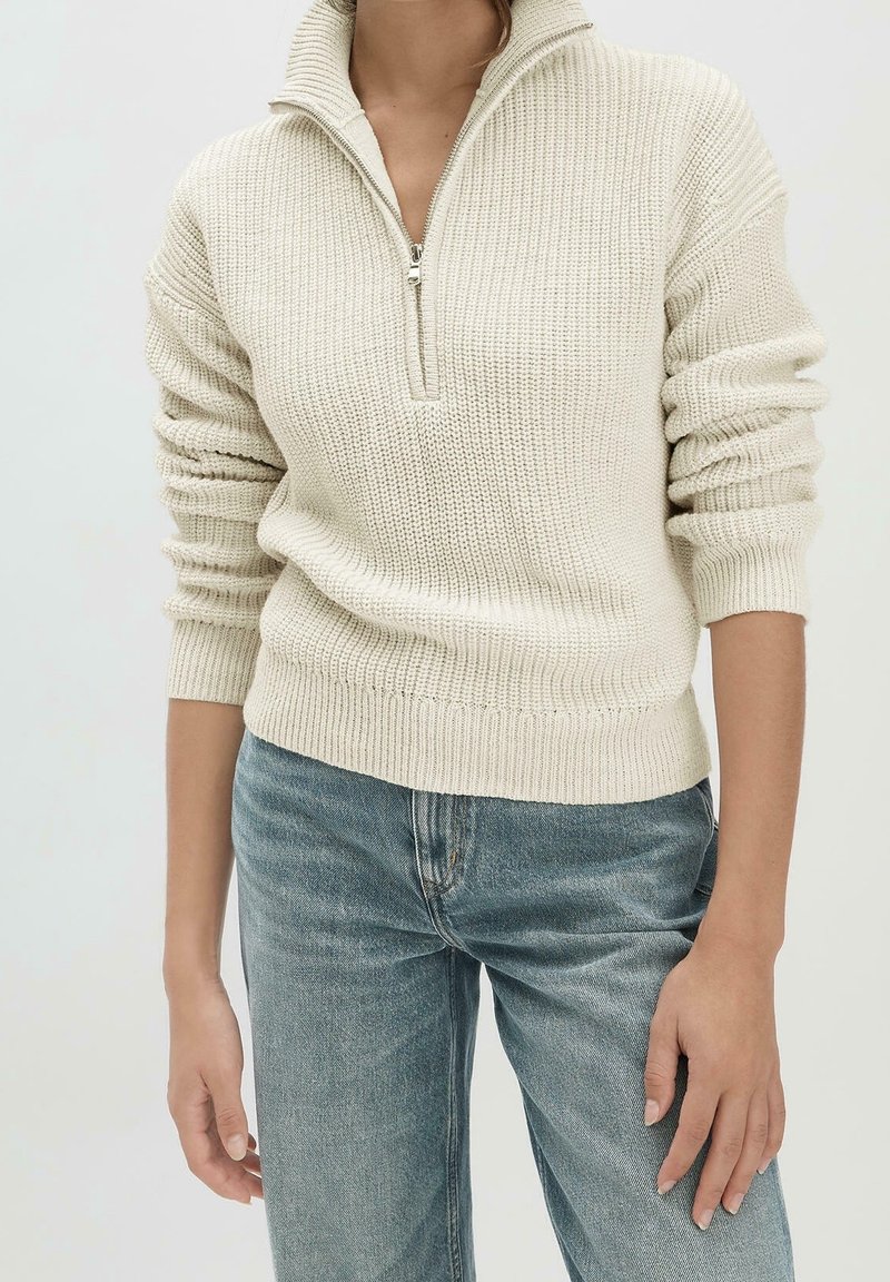 Frau, die einen cremefarbenen Strickpullover mit einem halben Reißverschlusskragen und langen Ärmeln trägt, kombiniert mit hochgeschnittenen blauen Jeans.