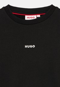 Svart hettegenser med ribbet rund hals, med en liten hvit "HUGO" logo på brystet og en rød detalj inni kragen.