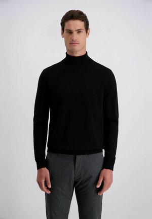 Uomo con un maglione nero a collo alto e pantaloni grigi, in piedi davanti a uno sfondo grigio chiaro uniforme.
