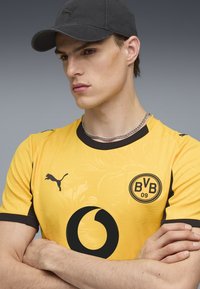 Geltona sportinė marškinė su juodais akcentais, turinti raštą, skydo logotipą ir Puma logotipą. Modelis dėvi juodą kepurę ir sidabrinę grandinėlę.
