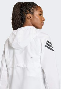 Biela nepremokavá bunda s kapucňou, ktorá má čierne pruhované akcenty na rukáve a ľahkú textúru. Zobrazený zadný pohľad.