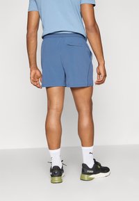 Pantaloni sportivi blu con tasca posteriore, vita elastica e dettagli riflettenti, abbinati a scarpe da corsa nere e calzini bianchi.