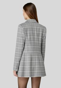 Blazer grigio a quadri con vestibilità sartoriale, chiusura a un bottone e dettagli con quattro bottoni sulle maniche. Tessuto liscio con leggero lucido.