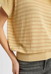 Krótki sweter z dzianiny w kolorze złotym z poziomymi paskami, z teksturowanym wzorem i ściągaczem na dole, zestawiony z ciemnoszarymi dżinsami.