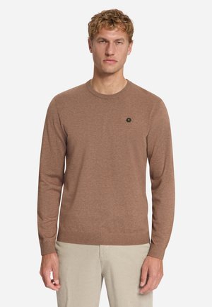 Homme blond portant un pull marron à manches longues col rond avec un petit logo sur la poitrine et un pantalon beige clair, debout face à l’avant.