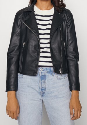 Persona con una chaqueta negra de cuero con cremallera, jersey de rayas blancas y negras, y vaqueros de tiro alto de color azul claro con las manos relajadas a los lados.