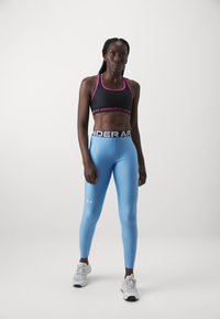 Črna športna modrc z roza akcenti, v kombinaciji s slim-fit svetlo modrimi legginsy in belimi supergami, z blagovno znamko Under Armour na pasu in nogi.