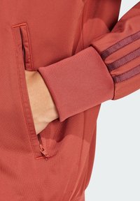 Chaqueta deportiva roja de corte ajustado, con tejido texturizado, bolsillo lateral con cremallera y franjas de acento marrón en las mangas.