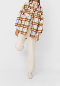 Chaqueta de camisa de cuadros en tonos crema, rojo, azul y amarillo, hecha de una tela texturizada con dos bolsillos en el pecho y detalles de botones. Pantalones de pierna ancha en color beige.