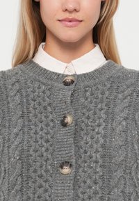 Grijze gebreide cardigan met een gestructureerd patroon, voorzien van grote ronde knopen en een geribbelde halslijn, gelaagd over een lichtroze overhemd met kraag.