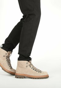 Piernas vestidas con pantalones negros y botas de senderismo de gamuza beige con ojales de metal y suelas de goma gruesa sobre un fondo blanco.