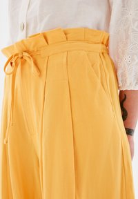 Gula linnbyxor med hög midja, knytdetalj och veck fram, kombinerat med en vit broderad blus.