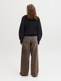 Woman with curly brown hair wearing a black sweater and loose brown pants, standing facing away on a plain white background.