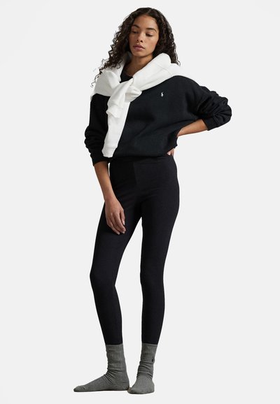 Černá mikina nošená s bílým svetr na zavazování, černými legínami a šedými žebrovanými ponožkami. Mikina má malé logo.