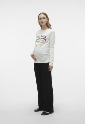 Långärmad krämfärgad gravid topp med julinspirerat text och röda och gröna detaljer, tillsammans med svarta vida byxor och svarta ballerinaskor.