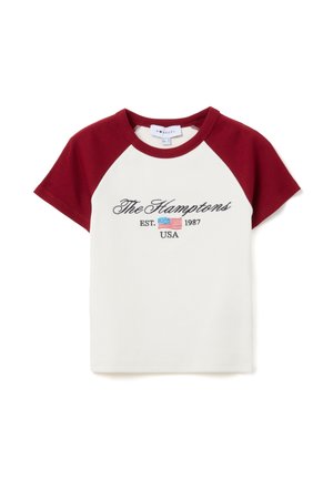 T-shirt blanc avec des manches raglan bordeaux. Présente un texte brodé "The Hamptons EST. 1987 USA" avec un petit motif de drapeau américain.