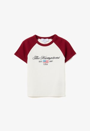 T-shirt blanc avec des manches raglan bordeaux. Présente un texte brodé "The Hamptons EST. 1987 USA" avec un petit motif de drapeau américain.