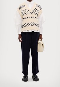 Gilet en tricot crème avec des motifs géométriques bleu marine, porté sur une chemise blanche à boutons. Un pantalon noir à jambes larges et un petit sac à main beige complètent la tenue.