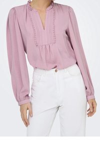 Blouse rose clair avec encolure à volants, plis et manches longues bouffantes, associée à un jean blanc taille haute. Tissu doux et texturé.