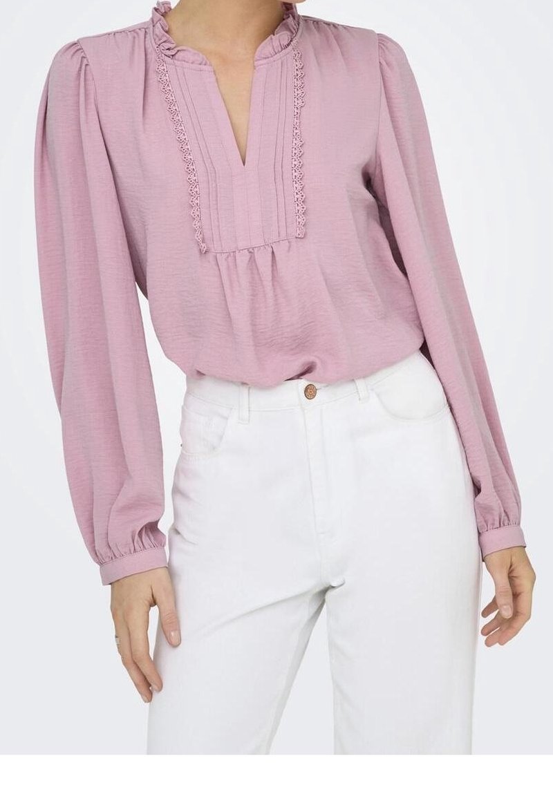 Blouse rose clair avec encolure à volants, plis et manches longues bouffantes, associée à un jean blanc taille haute. Tissu doux et texturé.