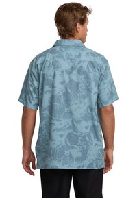 Man seen from the back wearing a short-sleeve, light blue patterned shirt and black pants against a white background.