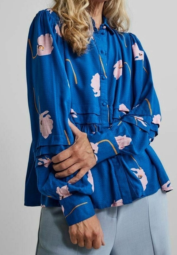 Femme portant un chemisier bleu à boutons avec un imprimé floral rose et des détails à volants, associée à un pantalon bleu clair, bras croisés devant elle.