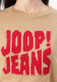 Beige gebreide trui met opvallende rode tekst "JOOP! JEANS." Ribbelkraag en een textuurdetail dat door de hele trui prominent aanwezig is.