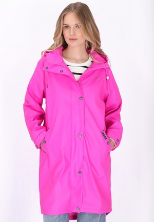 Nő élénk rózsaszín kapucnis esőkabátban, patentos gombokkal, csíkos ing fölött, kezét zsebekben tartva, fehér háttér előtt áll.