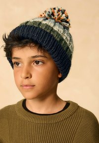 Bonnet en tricot bleu marine, vert et blanc avec un pompon coloré sur le dessus. L'enfant porte un pull texturé vert olive avec un col rond.
