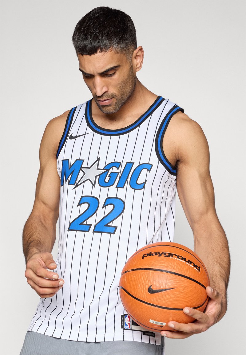 Weißes Basketballtrikot mit vertikalen schwarzen Streifen, blauen Akzenten, "MAGIC" und der Nummer "22" in Blau. Hält einen orangefarbenen Basketball.