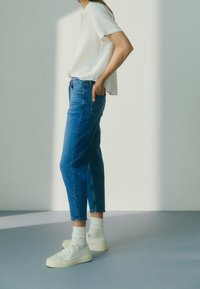 Modré džínové kalhoty se zužujícím střihem, bílé tričko a světlé tenisky. Džíny mají standardní kapsy a detail s neupravovaným lemem.