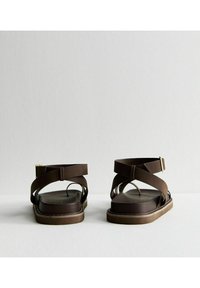 Sandali marroni con un ampio design a bande incrociate, suole piatte e una superficie testurizzata. Chiusura con fibbia regolabile alla caviglia.