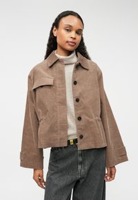 Chaqueta de pana marrón con cuello, cierre frontal de botones y bolsillo tipo parche. Presenta mangas anchas y un corte relajado.