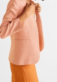 Person adjusting collar of a textured light pink blazer worn over an orange skirt or pants, against a plain white background.
