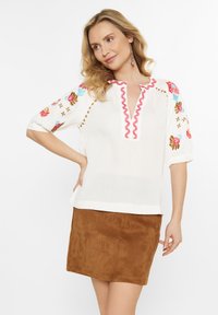 Bluză albă texturată cu broderie florală colorată pe mâneci, detaliu de guler ruffled roz, combinată cu o fustă din suede maro.