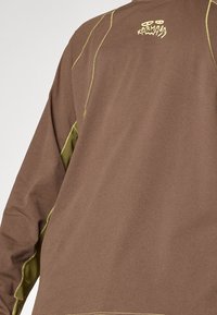 Brown long-sleeve shirt with green accents and stitching. Features a logo on the back and a smooth, textured fabric design.