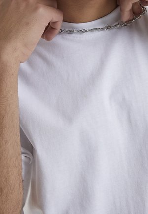 Hände halten eine verdrehte silberne Kettenhalskette vor einem weißen T-Shirt mit Rundhalsausschnitt.