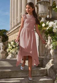 Világos rózsaszín ruha csipkebetéttel az felsőrészen, fodros ujjakkal és lenge szoknyával. Ezüst cipővel párosítva, fehér virágcsokrot tartva a kezében.