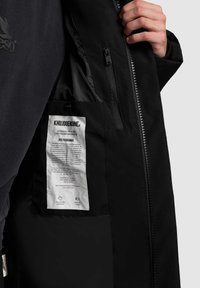 Juoda striukė su užtrauktuku, turi vidinę kišenę ir priežiūros etiketę. Medžiaga atrodo lygi su matinio paviršiaus efektu.