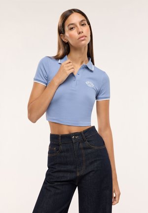 Giovane donna con polo corta blu e jeans scuri a vita alta, in piedi davanti a uno sfondo semplice, che posa con una mano vicino al colletto.