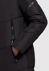 Sekcja czarnej kurtki puchowej zbliżenie na rękaw z kieszonką na zamek. Tkanina jest gładka, a projekt obejmuje pikowane panele.