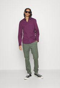 Camicia viola con bottoni e maniche lunghe, abbinata a pantaloni verdi. Sneakers nere con suole e lacci bianchi. Il modello indossa occhiali da sole.