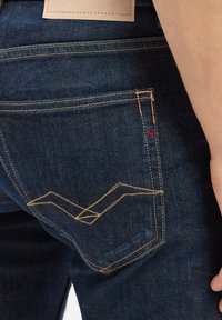 Mörkblå denimjeans har kontrasterande gula sömmar och ett unikt zickzackmönster på bakfickan. Läderetikett ovanför midjebandet.