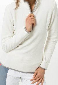 Persona ajustando la cremallera de un pulóver de forro blanco, vistiendo pantalones blancos con un parche gris y rojo.