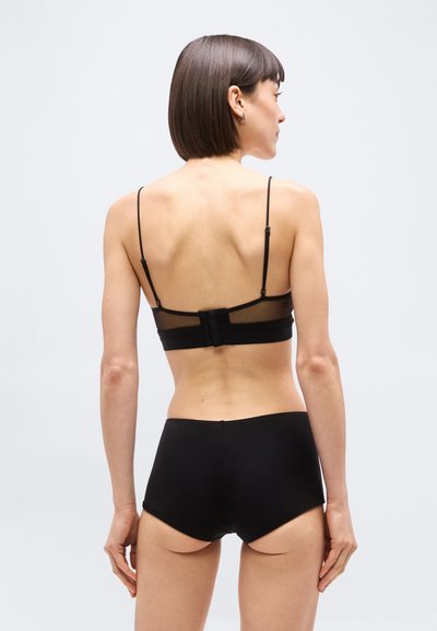Γυναίκα με ίσια σκούρα μαλλιά που φοράει μαύρο σουτιέν με ρυθμιζόμενες τιράντες και μαύρο εσώρουχο με ψηλή μέση, στέκεται μπροστά σε απλό λευκό φόντο.